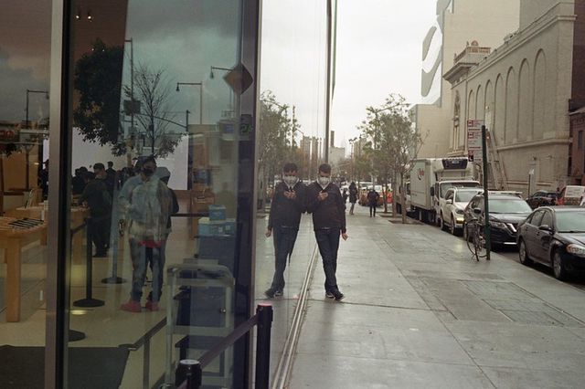 A man wearing a mask leans against a glass storefront and his reflection while staring at his phone.